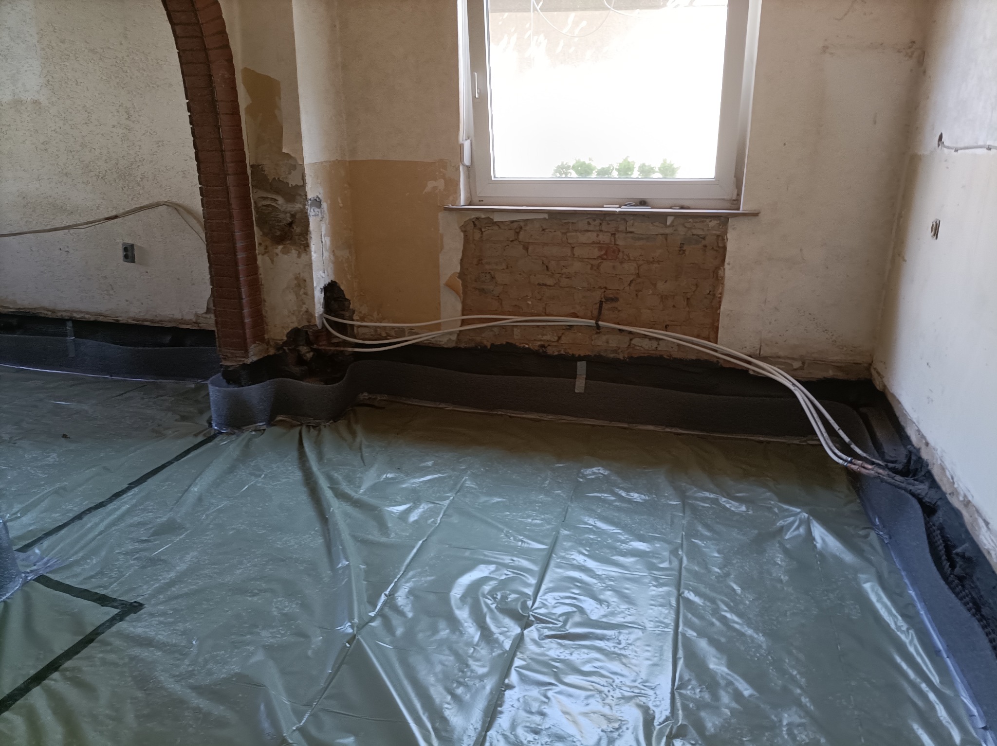 Przygotowanie podłoża: folia ochronna, rury grzewcze i izolacja pionowa przy ścianach w surowym wnętrzu z oknem i fragmentem ceglanej ściany.