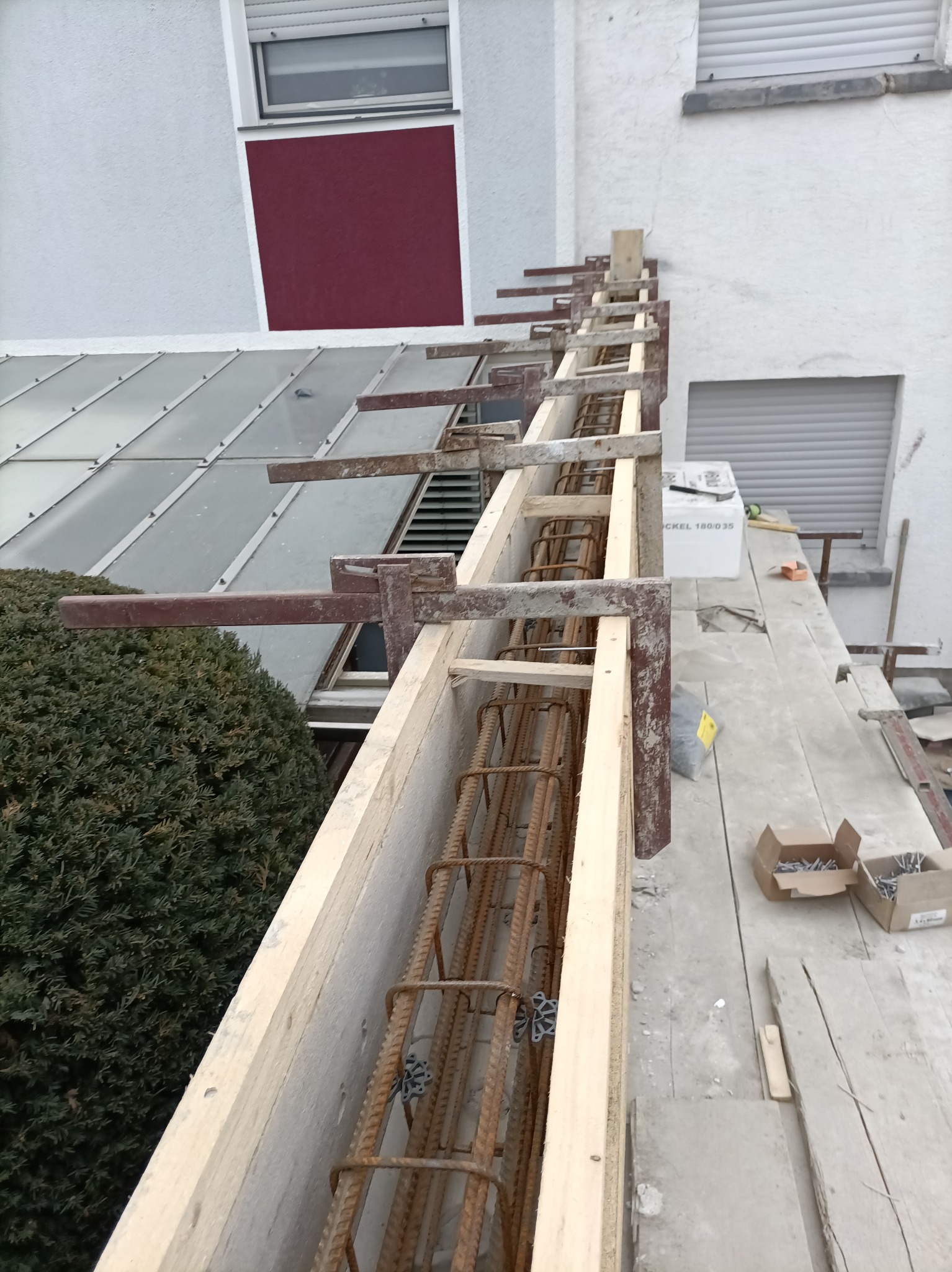 Szczegółowe ujęcie z góry na zbrojenie w drewnianym szalunku w trakcie budowy, z widocznymi elementami mocującymi i fragmentem elewacji budynku w tle.