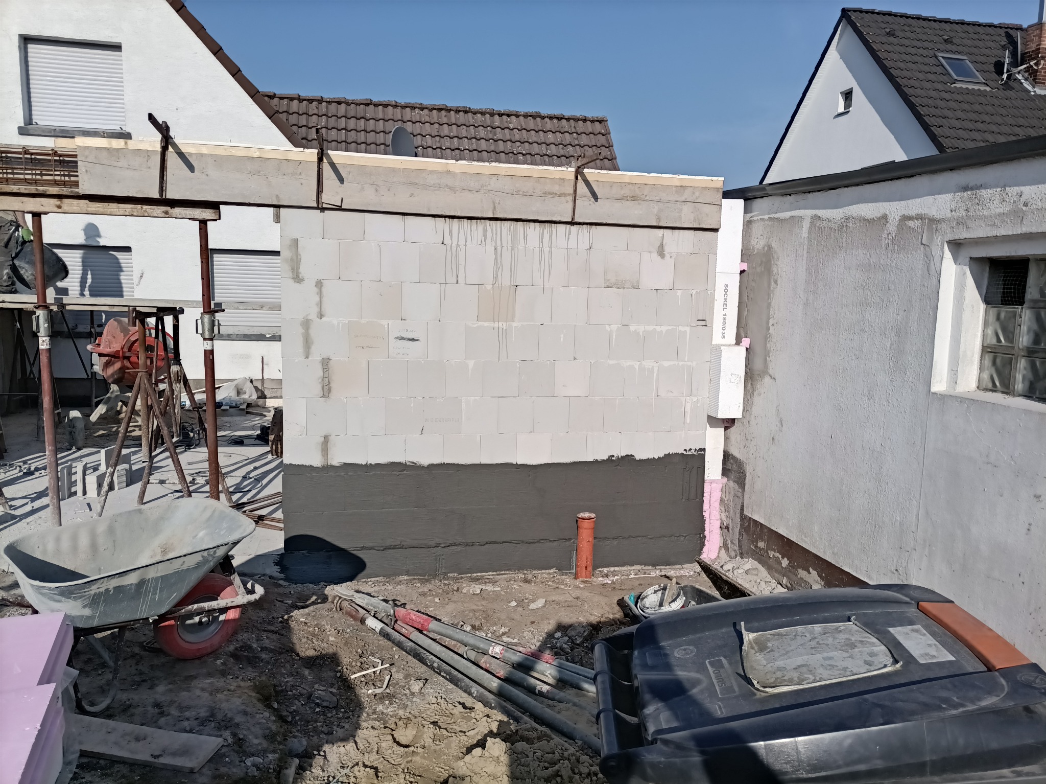 Wznoszenie ściany z bloczków, z widoczną izolacją fundamentów i elementami konstrukcyjnymi. Na pierwszym planie taczka i gruz, w tle budynki mieszkalne.