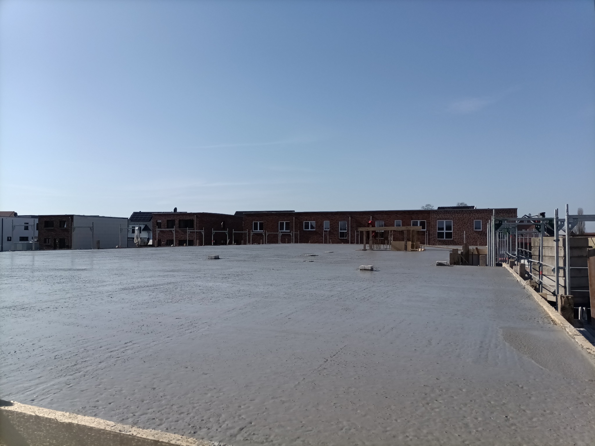 Wylany beton na dużej powierzchni budowy, z widocznymi budynkami w tle i niebieskim niebem. Widoczne elementy konstrukcyjne i rusztowania.