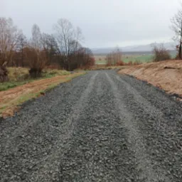 Świeżo utwardzona droga szutrowa z widocznymi śladami po przejeździe, otoczona ziemią i rzadką roślinnością, w tle zalesione wzgórza pod pochmurnym niebem.