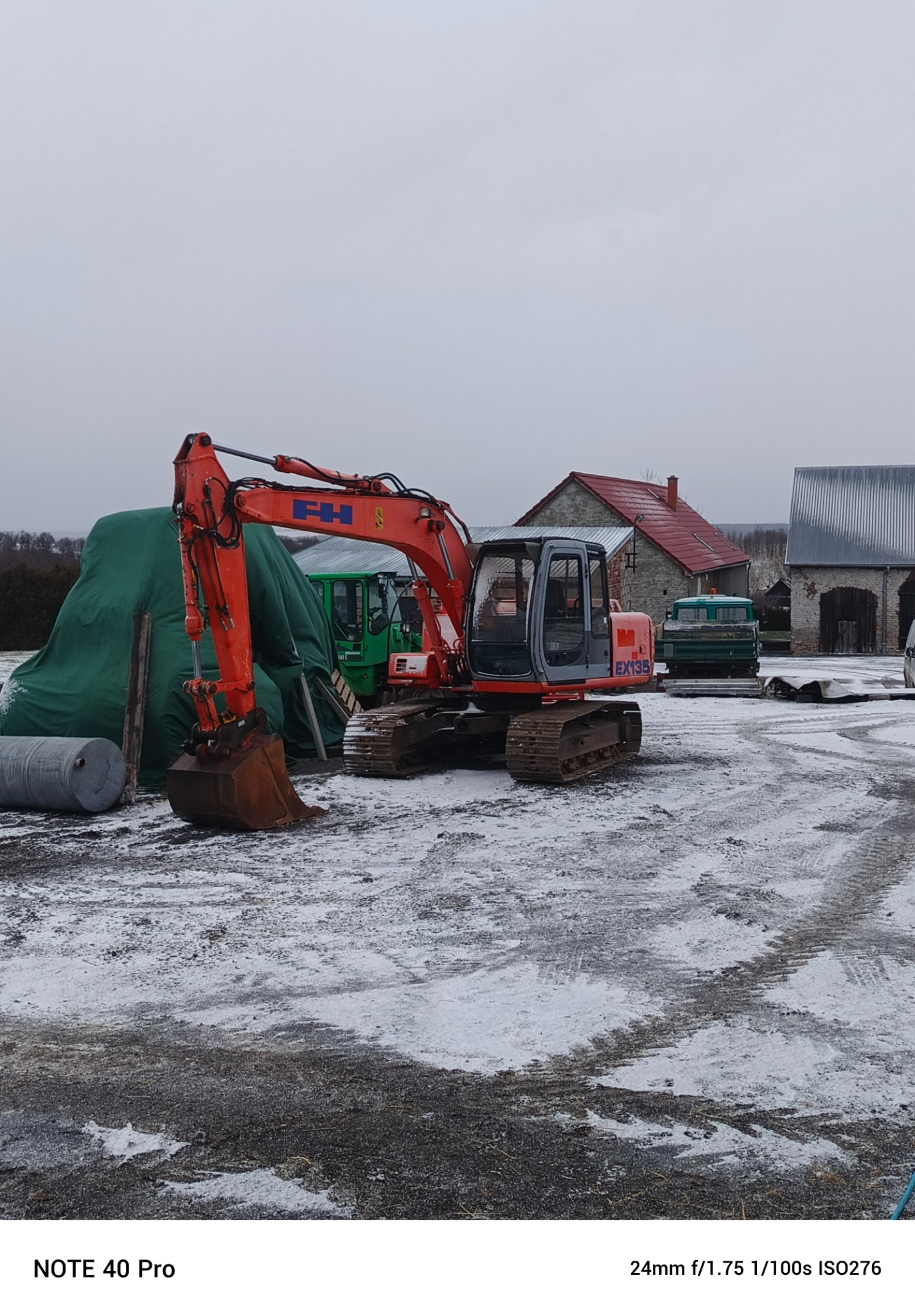 Pomarańczowa koparka EX135 na placu pokrytym śniegiem, w tle budynki gospodarcze i zielony traktor pod plandeką.