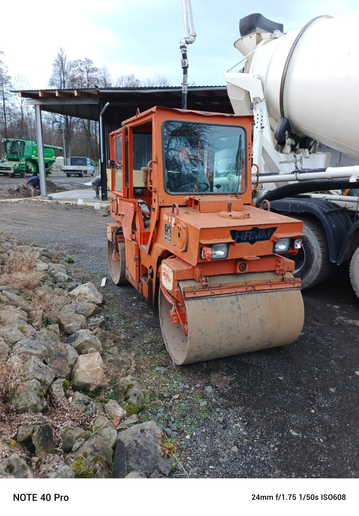 Pomarańczowy walec drogowy HAMM DV 3 w Mirsku stoi na żwirowym podłożu obok betonomieszarki, w tle widoczny budynek gospodarczy.