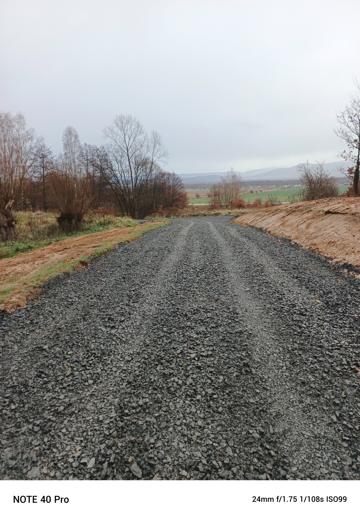 Świeżo utwardzona droga szutrowa z widocznymi śladami po przejeździe, otoczona ziemią i rzadką roślinnością, w tle zalesione wzgórza pod pochmurnym niebem.