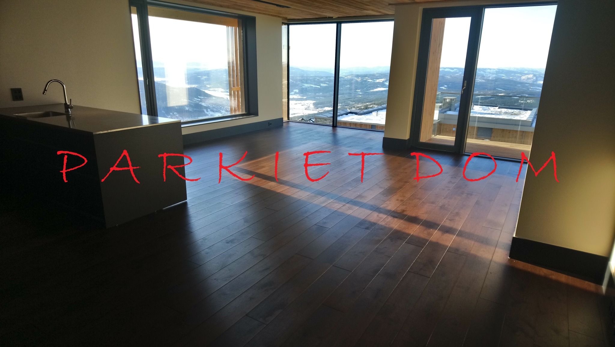 Ciemny, lakierowany parkiet w przestronnym salonie z widokiem na góry, duże okna z drewnianymi ramami, minimalistyczna wyspa kuchenna z chromowaną baterią.