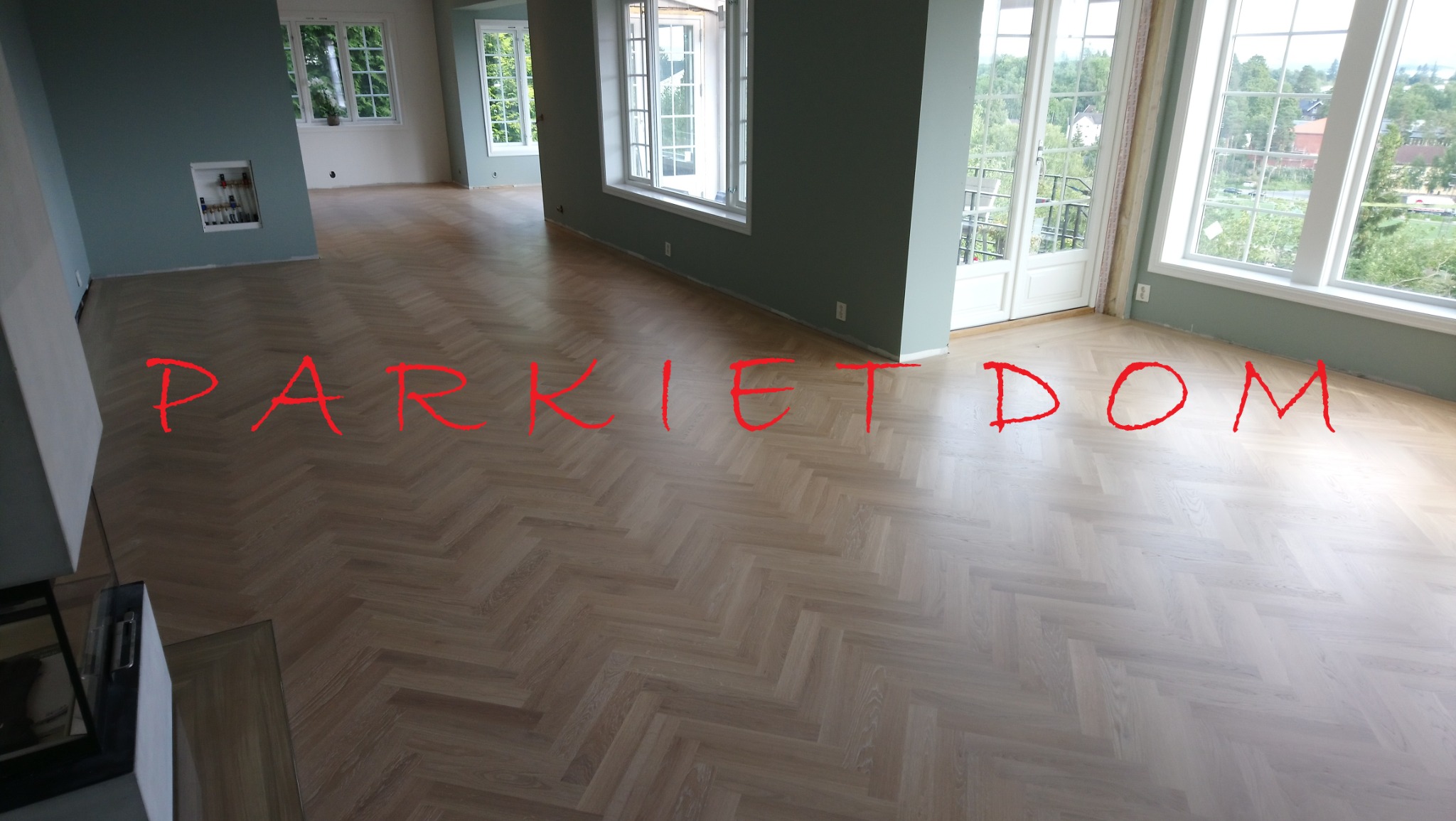 Jasny parkiet ułożony we wzór jodełki francuskiej w przestronnym, słonecznym salonie z dużymi oknami i zielonym widokiem na zewnątrz.