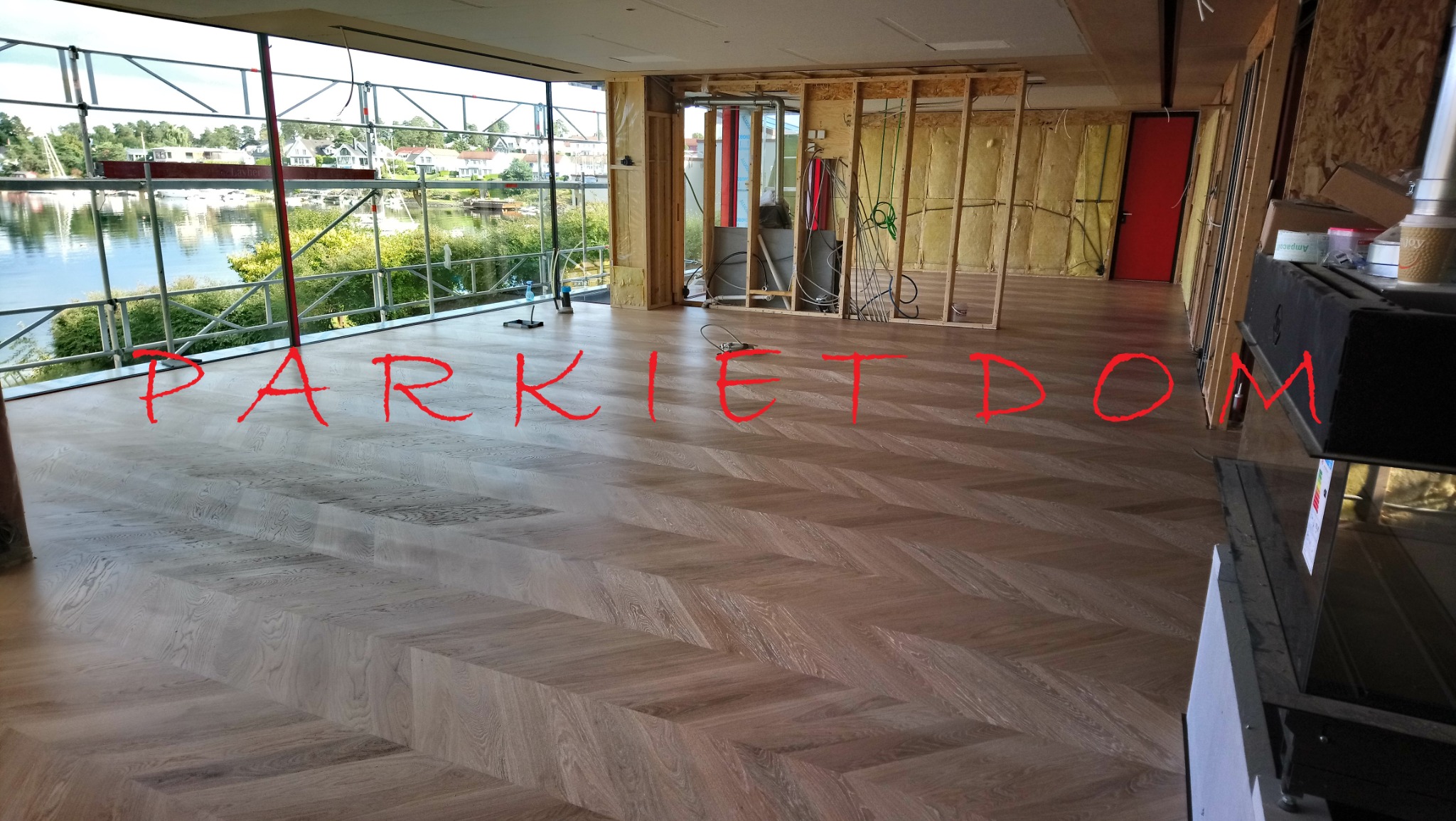 Nowo ułożona podłoga z parkietu w jodełkę francuską w przestronnym wnętrzu w trakcie wykańczania, z widokiem na jezioro za oknem.