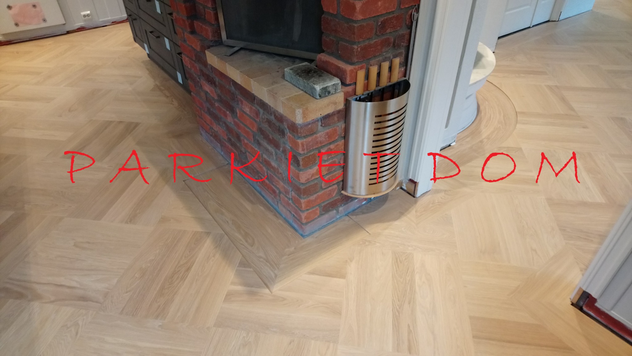 Ułożony parkiet w jodełkę francuską w Kielcach, otaczający ceglany kominek z metalowym pojemnikiem na drewno, widoczne dopasowanie parkietu do futryny drzwi.