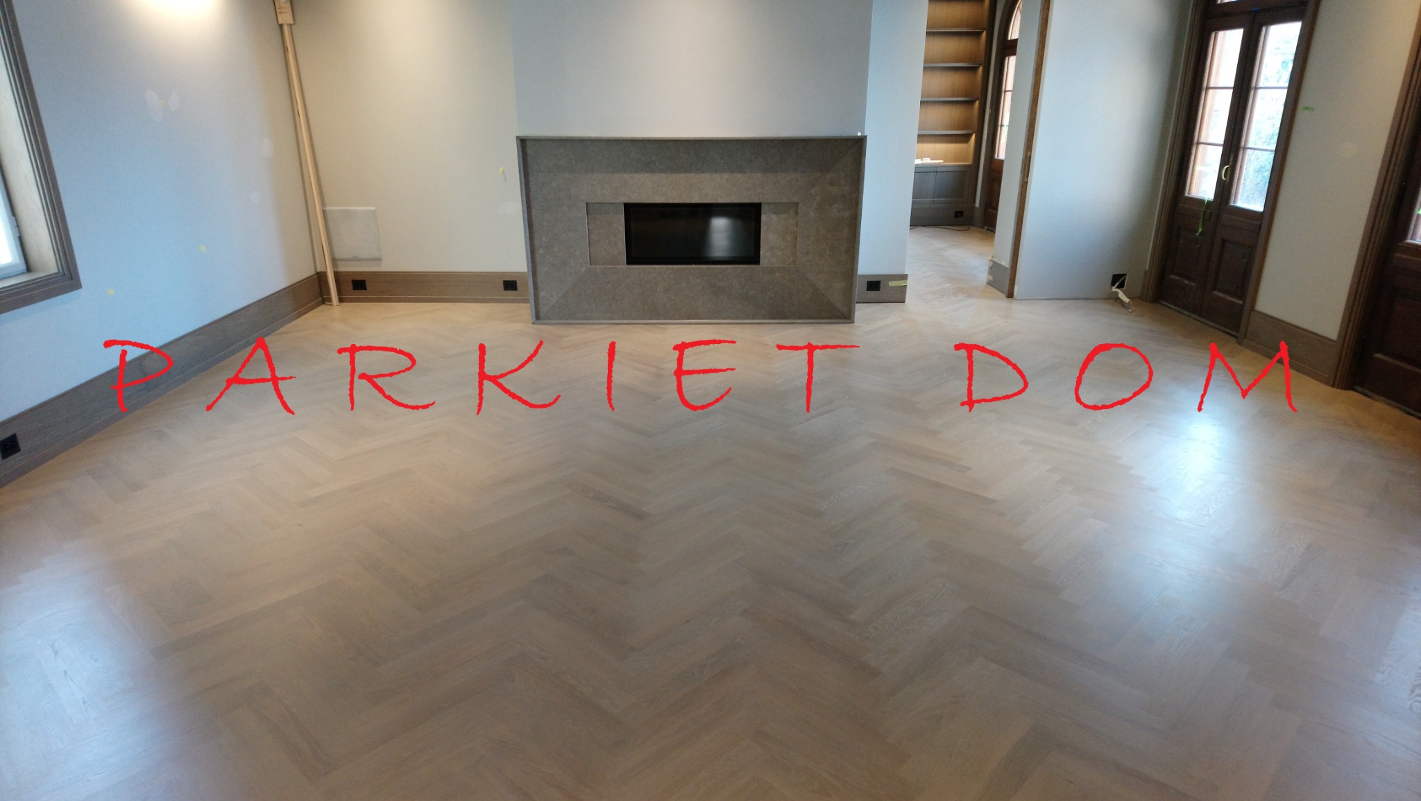 Jasny parkiet ułożony we wzór jodełki francuskiej w przestronnym salonie z kominkiem i drewnianymi drzwiami.