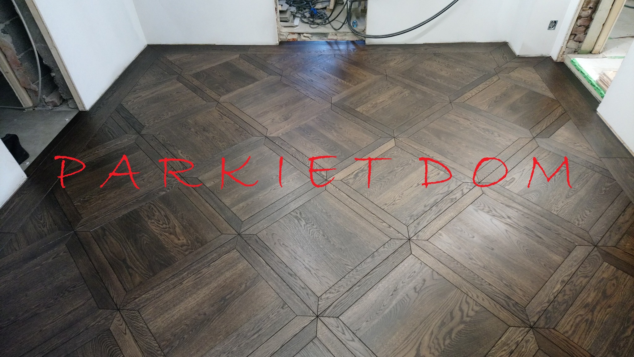 Ukończona podłoga z ciemnego parkietu ułożonego we wzór rombów w trakcie remontu, widoczne kable i niedokończone ściany.