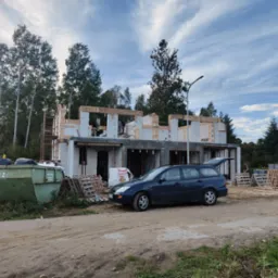 Wznoszenie surowego domu z białych bloczków, widoczne drewniane szalunki, pracownicy na rusztowaniu, samochód osobowy z otwartą klapą bagażnika i kontener na odpady budowlane z numerem 12.