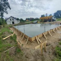 Wylany fundament pod budowę domu jednorodzinnego z drewnianym szalunkiem, widoczny betonowóz z pompą i częściowo ukończony budynek w tle.