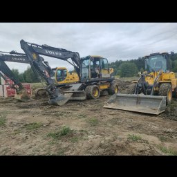 INVEST ELITE - Żółte maszyny budowlane Volvo: koparka kołowa z numerem 663 694 7 i ładowarka kołowa z łyżką na placu budowy.