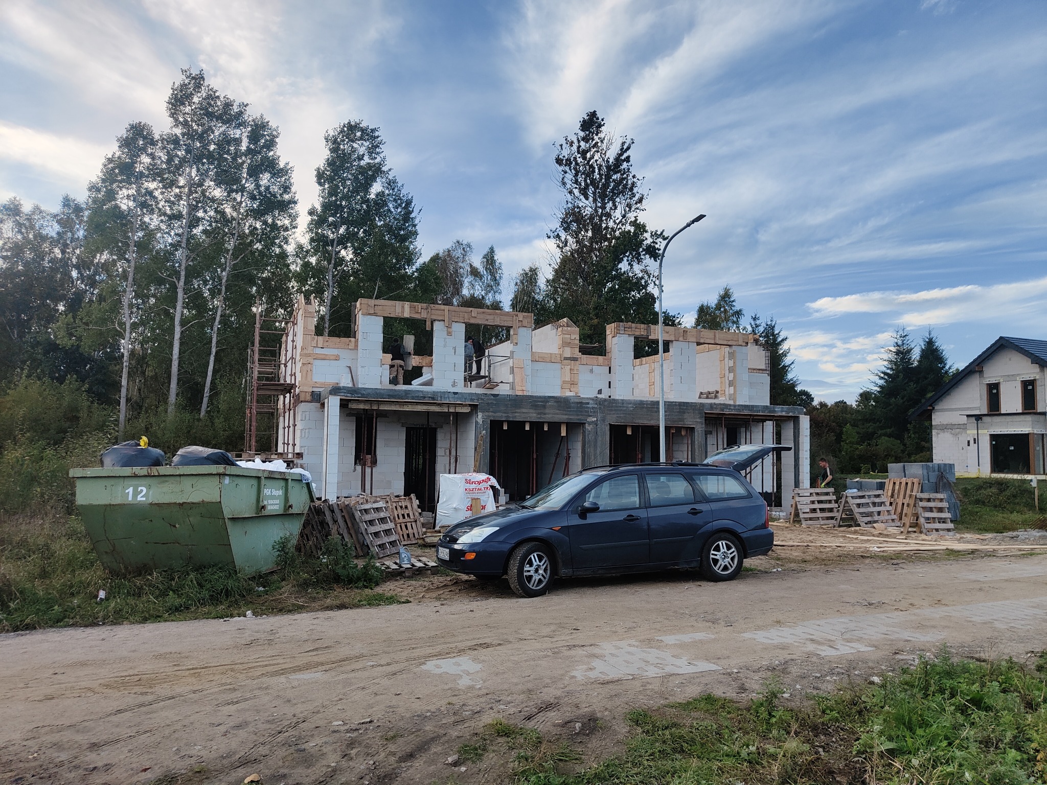 Wznoszenie surowego domu z białych bloczków, widoczne drewniane szalunki, pracownicy na rusztowaniu, samochód osobowy z otwartą klapą bagażnika i kontener na odpady budowlane z numerem 12.