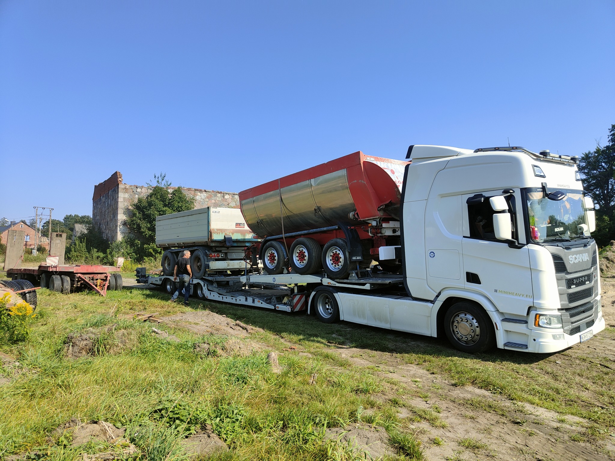 Biała ciężarówka SCANIA z naczepą niskopodwoziową przewożąca dwie przyczepy wywrotki w terenie trawiastym, w tle ruiny budynku i błękitne niebo.