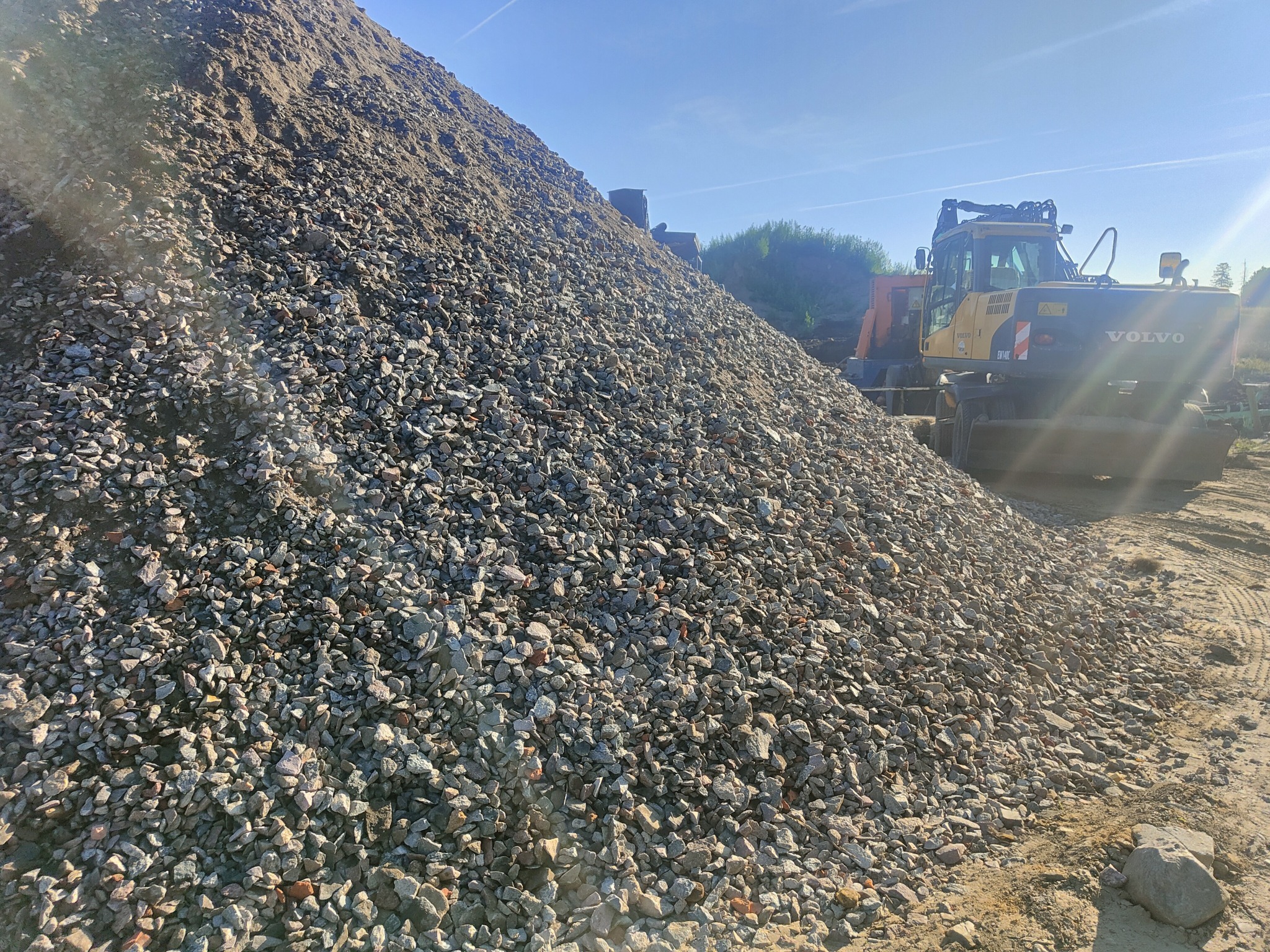 Góra szarego kruszywa z widoczną w tle żółto-czarną koparką kołową Volvo EW140C w słoneczny dzień.