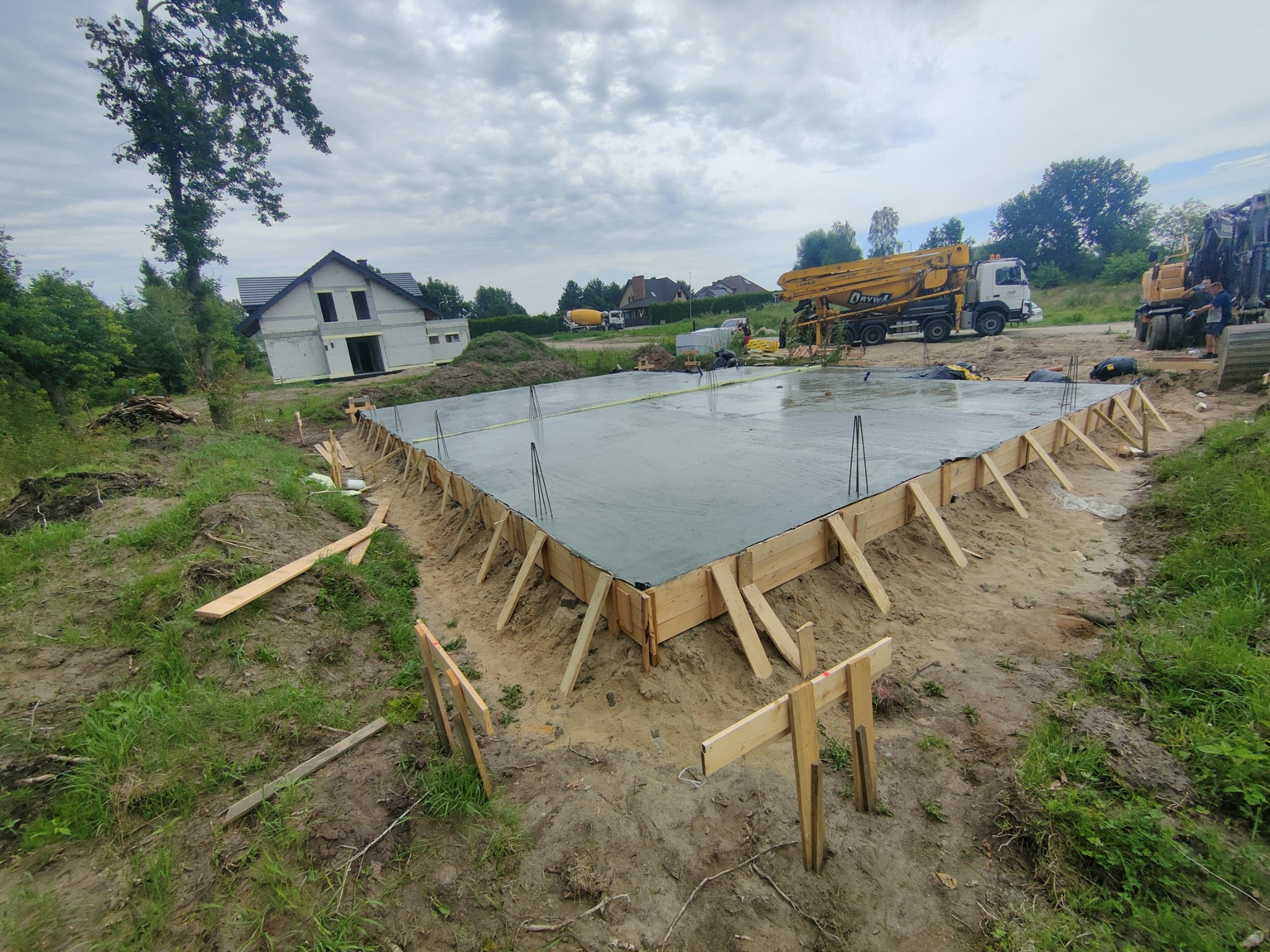 Wylany fundament pod budowę domu jednorodzinnego z drewnianym szalunkiem, widoczny betonowóz z pompą i częściowo ukończony budynek w tle.
