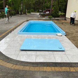 Krystian Chojak Bruk - Montaż basenu ogrodowego: ułożona kostka brukowa w łuku, płytki wokół basenu, przygotowany teren pod dalsze prace. Widoczni pracownicy i dom w tle.