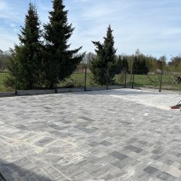 Krystian Chojak Bruk - Ułożona, szara kostka brukowa z fugami zasypanymi piaskiem, czyszczona szczotką w Ciepielowie. Widoczne drzewa iglaste i ogrodzenie.
