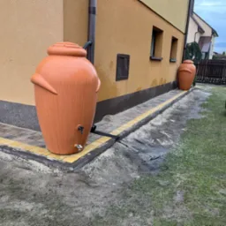 Pomarańczowe zbiorniki na deszczówkę w kształcie amfor, ustawione na obrzeżach z kostki brukowej wzdłuż ściany budynku. W tle widoczny pies i żółty pojemnik.