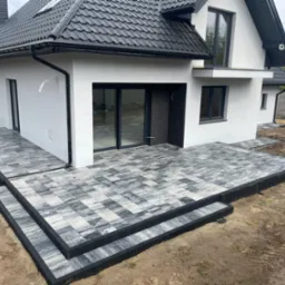 Wykończony taras z kostki brukowej w odcieniach szarości, z czarnym obrzeżem, przylegający do nowoczesnego, białego domu z czarnym dachem i stolarką okienną.