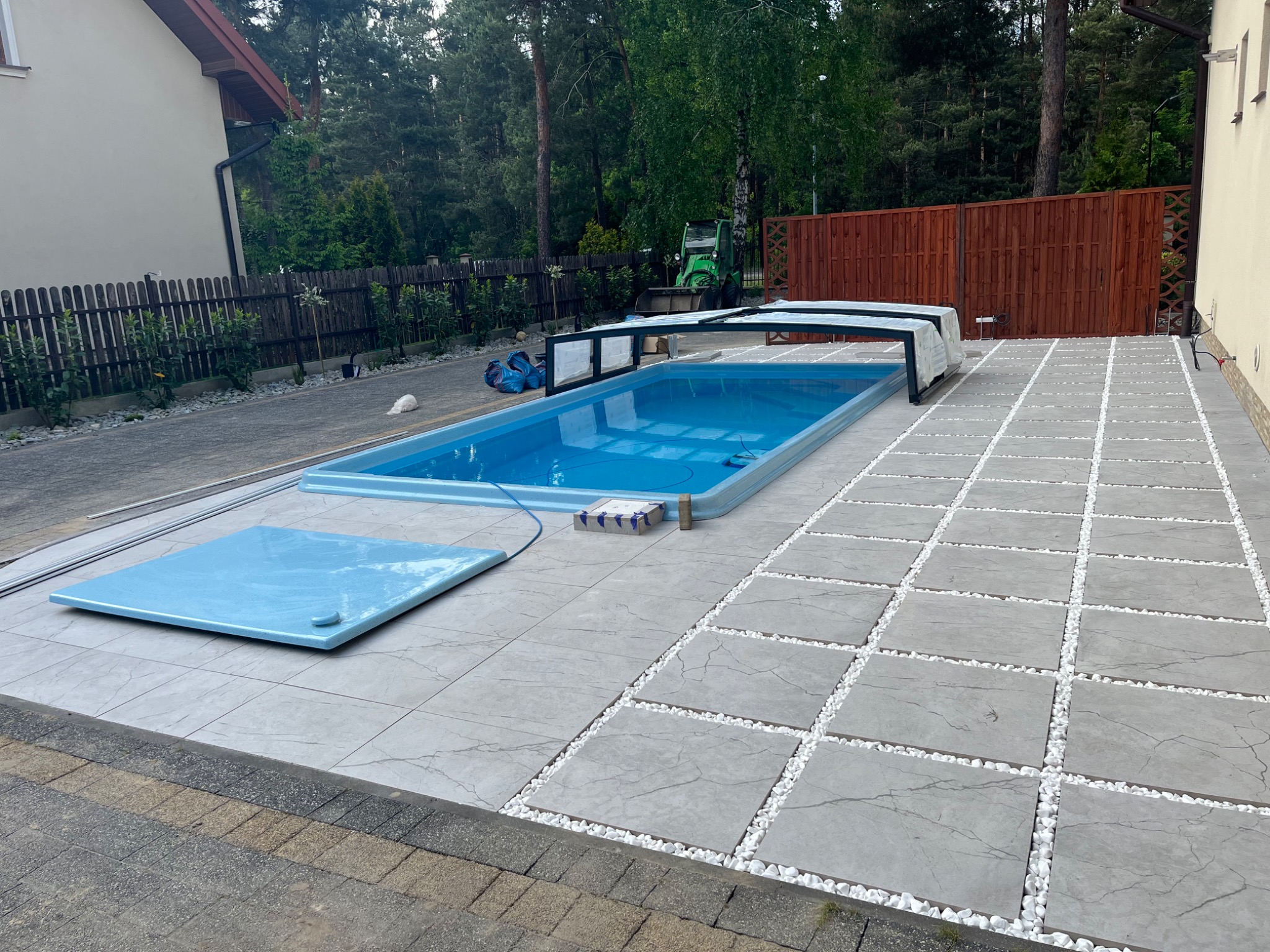 Nowoczesny basen z szarą kostką i białymi kamykami, otoczony zielenią i ogrodzeniem. Widoczny fragment domu i maszyna budowlana w tle.