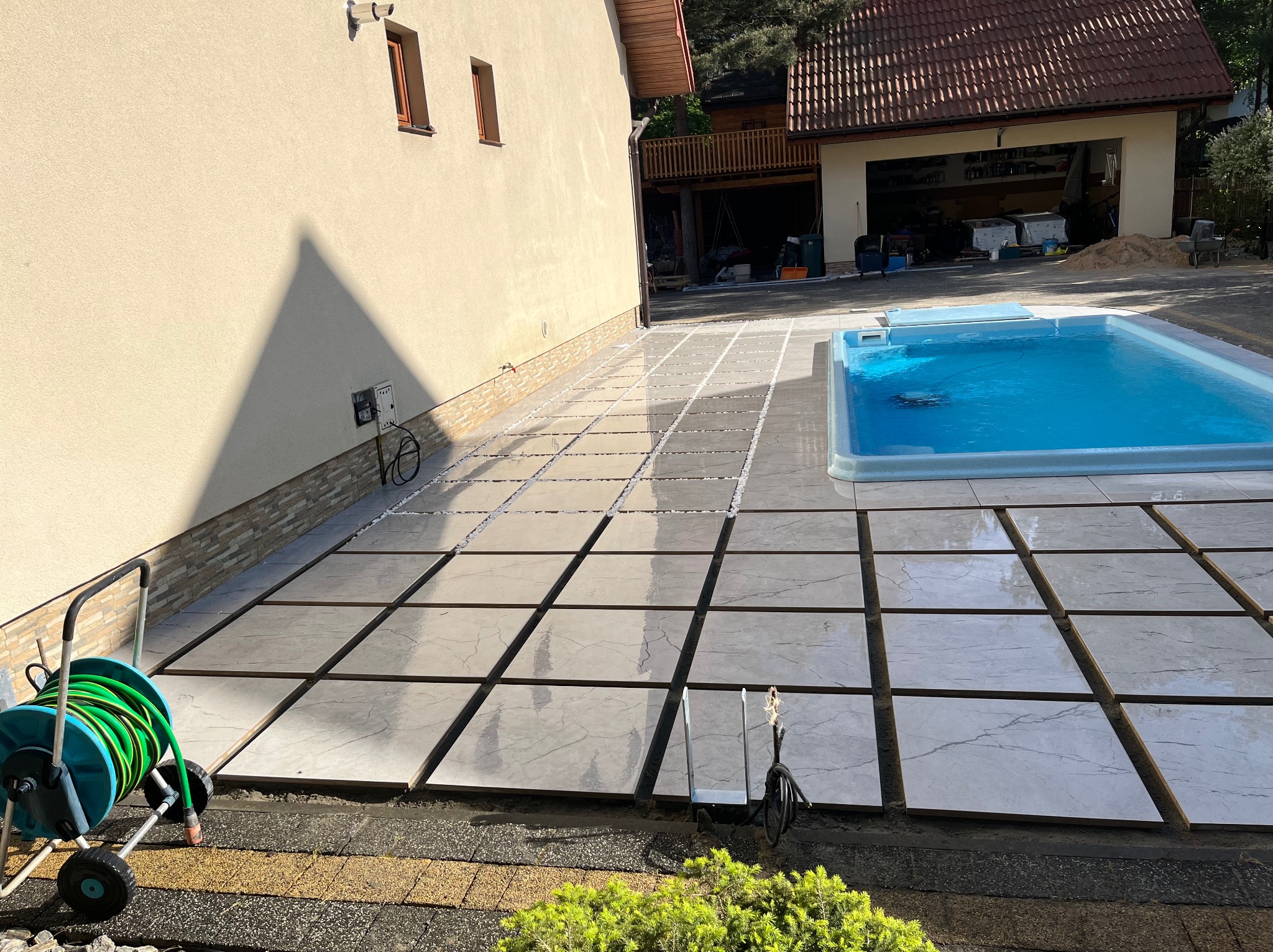 Układanie płyt tarasowych wokół basenu. Widoczne szczeliny wypełnione białym żwirem, zieleń i zwinięty wąż ogrodowy. Częściowo ułożona nawierzchnia.