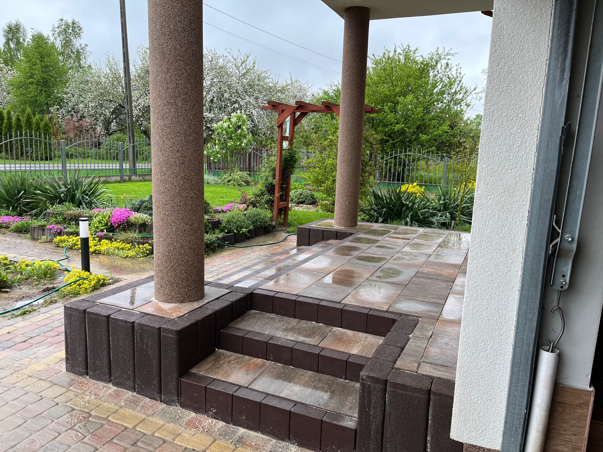 Zadaszony taras z kostki brukowej w odcieniach brązu i beżu, z kolumnami i schodami. W tle ogród z kwitnącymi drzewami i krzewami.