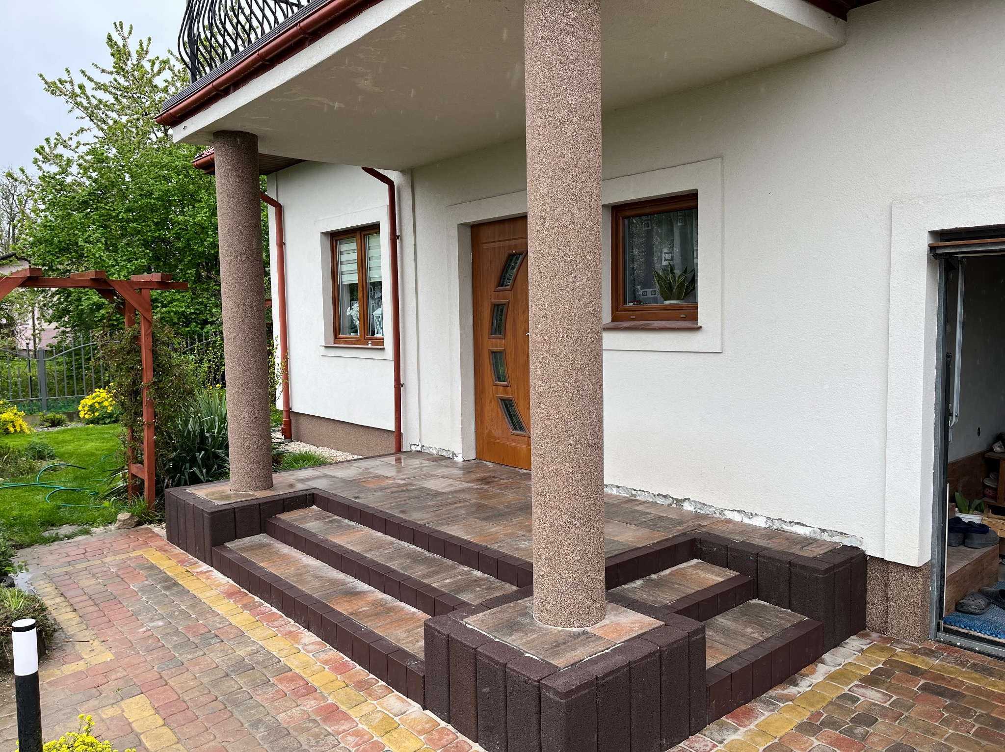 Nowoczesne schody wejściowe z kostki brukowej w odcieniach brązu i beżu, prowadzące do domu z kolumnami. Widoczny detal kolorowej kostki na chodniku.