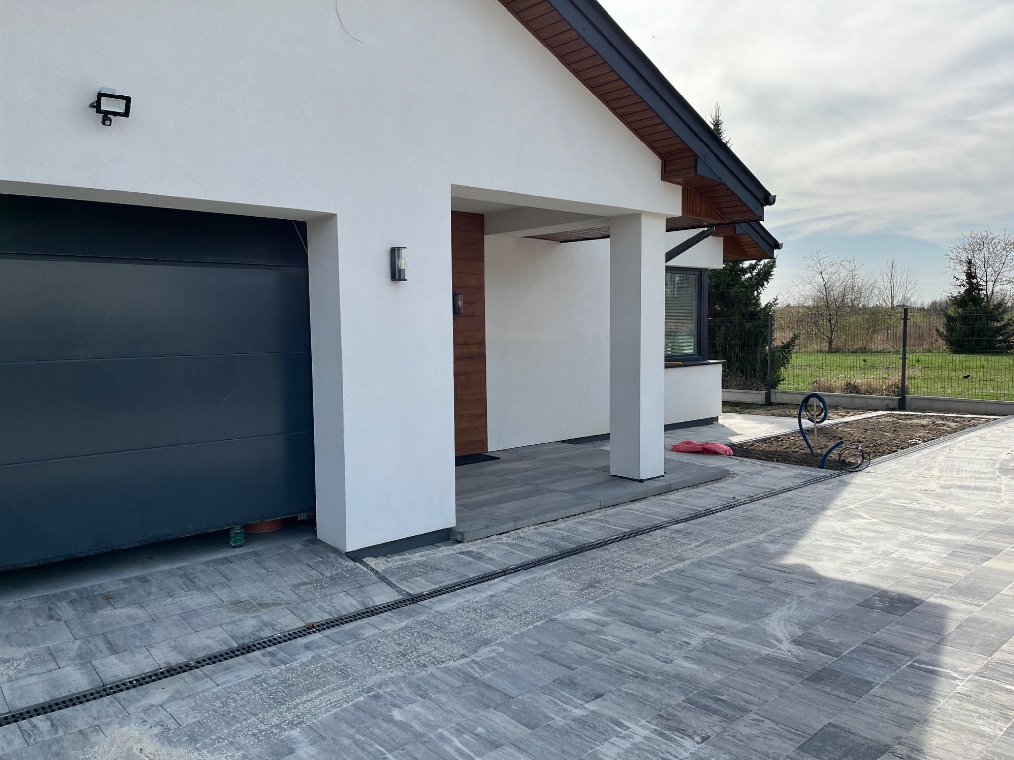 Nowoczesny dom z szarą kostką brukową. Widoczny garaż, kolumny podtrzymujące dach i rabata z elementami dekoracyjnymi. Ujęcie z poziomu gruntu, w słoneczny dzień.