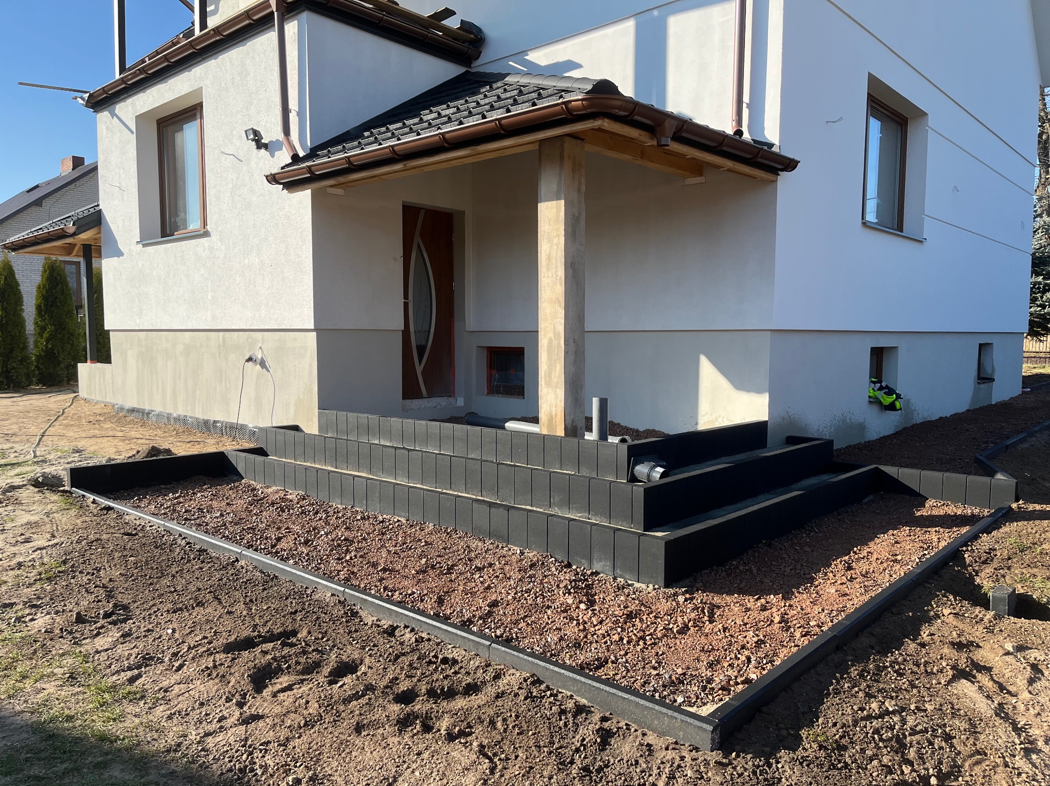 Nowoczesne schody wejściowe z czarnej kostki brukowej prowadzące do domu z białym tynkiem, otoczone czerwoną żwirową podsypką. Widok na elewację i fragment ogrodu.