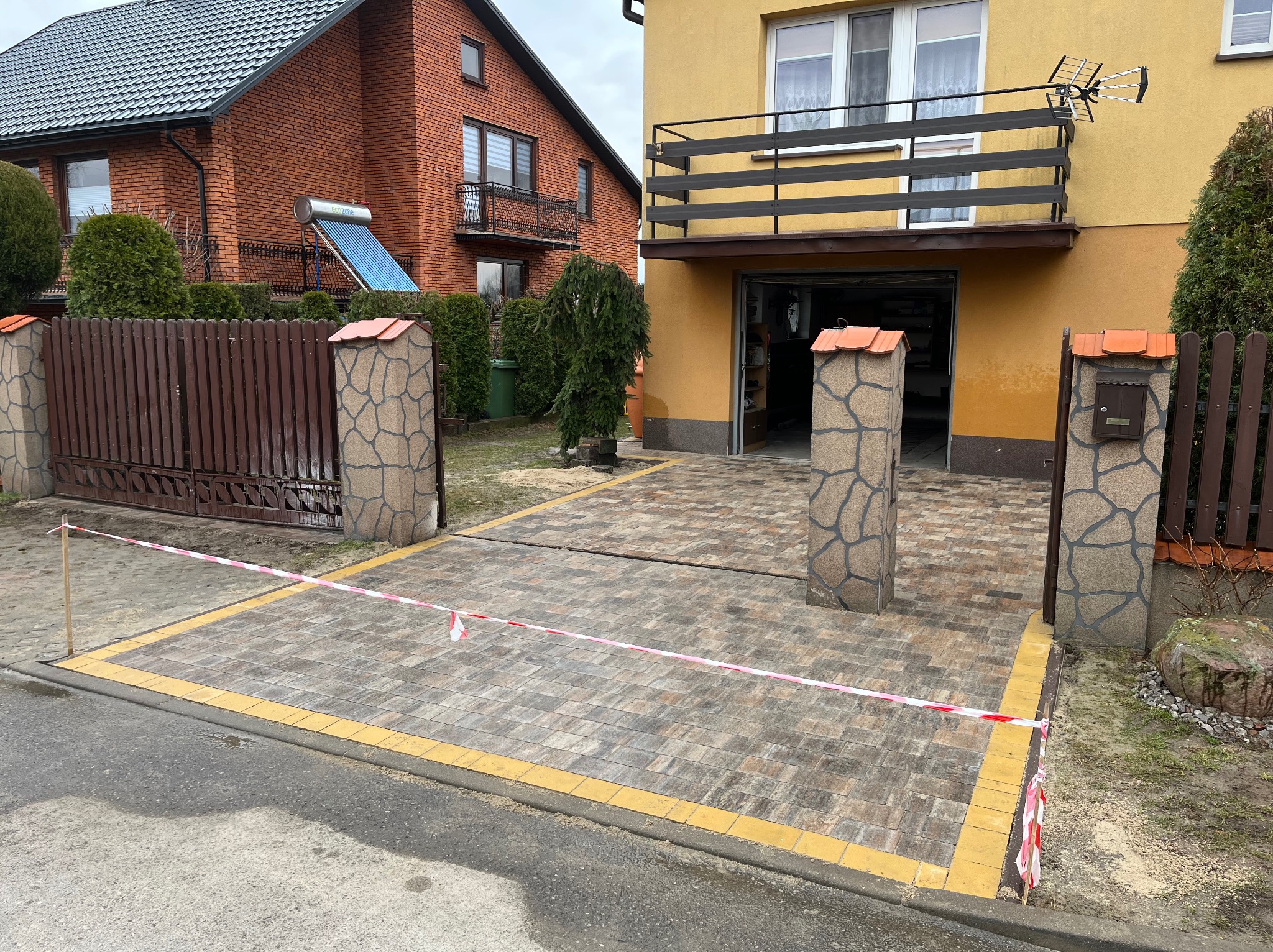Nowa, beżowo-szara kostka brukowa z żółtymi obrzeżami przed posesją z dwoma budynkami, oddzielona taśmą ostrzegawczą. Widoczne słupki ogrodzeniowe.