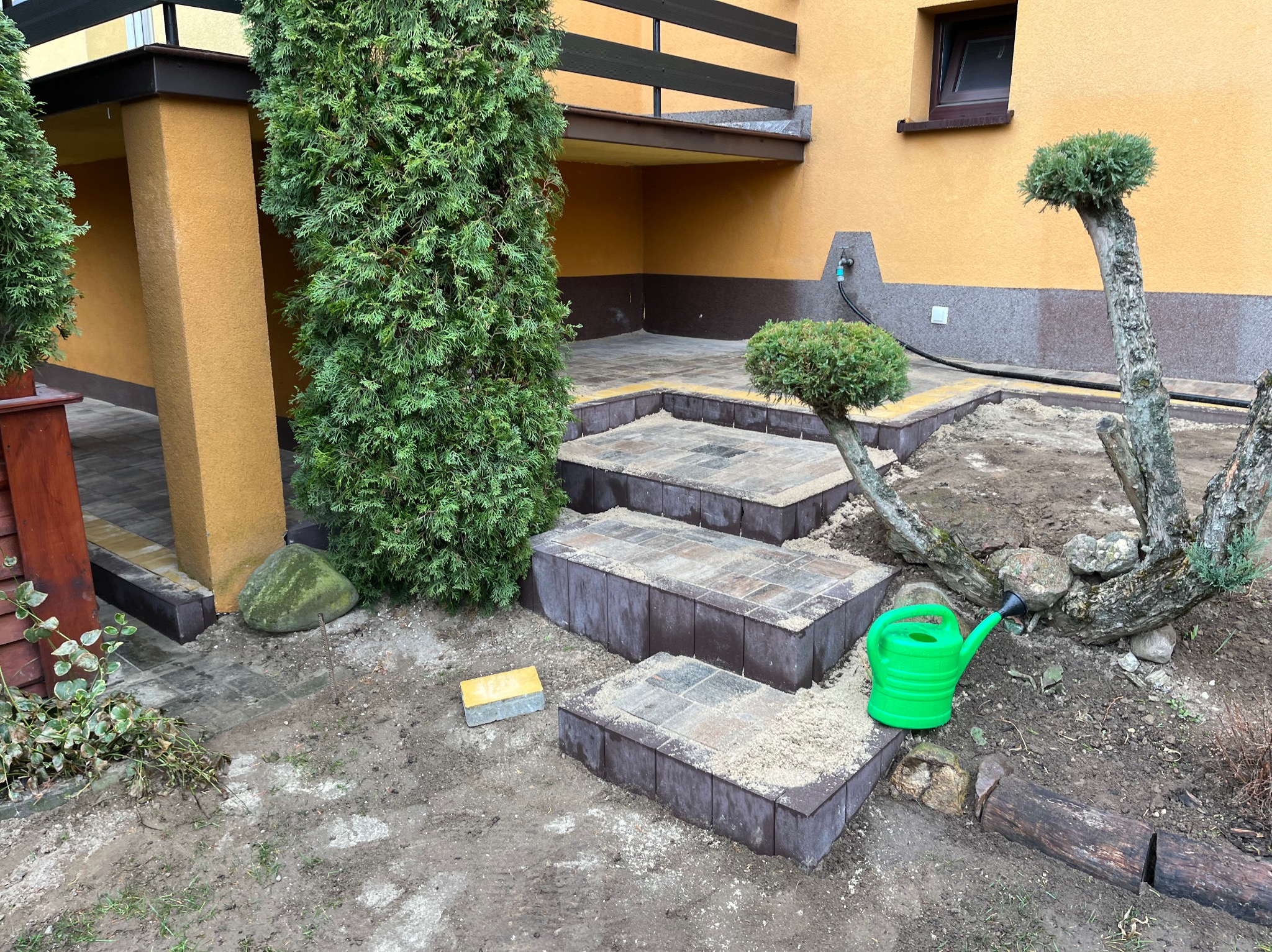 Kaskadowe schody z kostki brukowej w ogrodzie, w trakcie budowy. Widoczne elementy krajobrazu: drzewa, krzewy, trawnik. W tle żółta elewacja budynku.