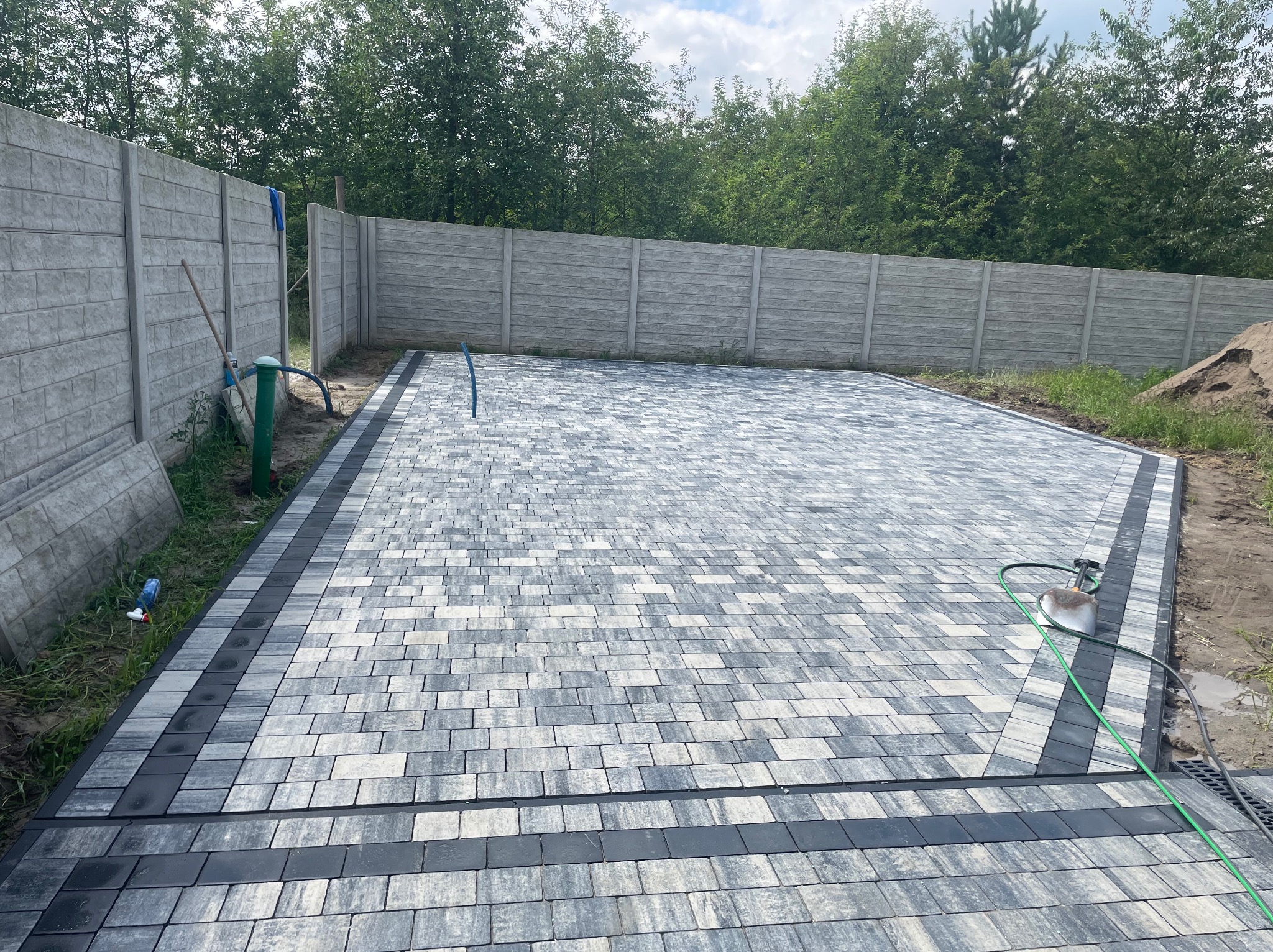Ułożona szara kostka brukowa z ciemnymi obrzeżami na posesji z betonowym ogrodzeniem. Widoczny wąż ogrodowy, drzewa w tle i fragment sterty ziemi.