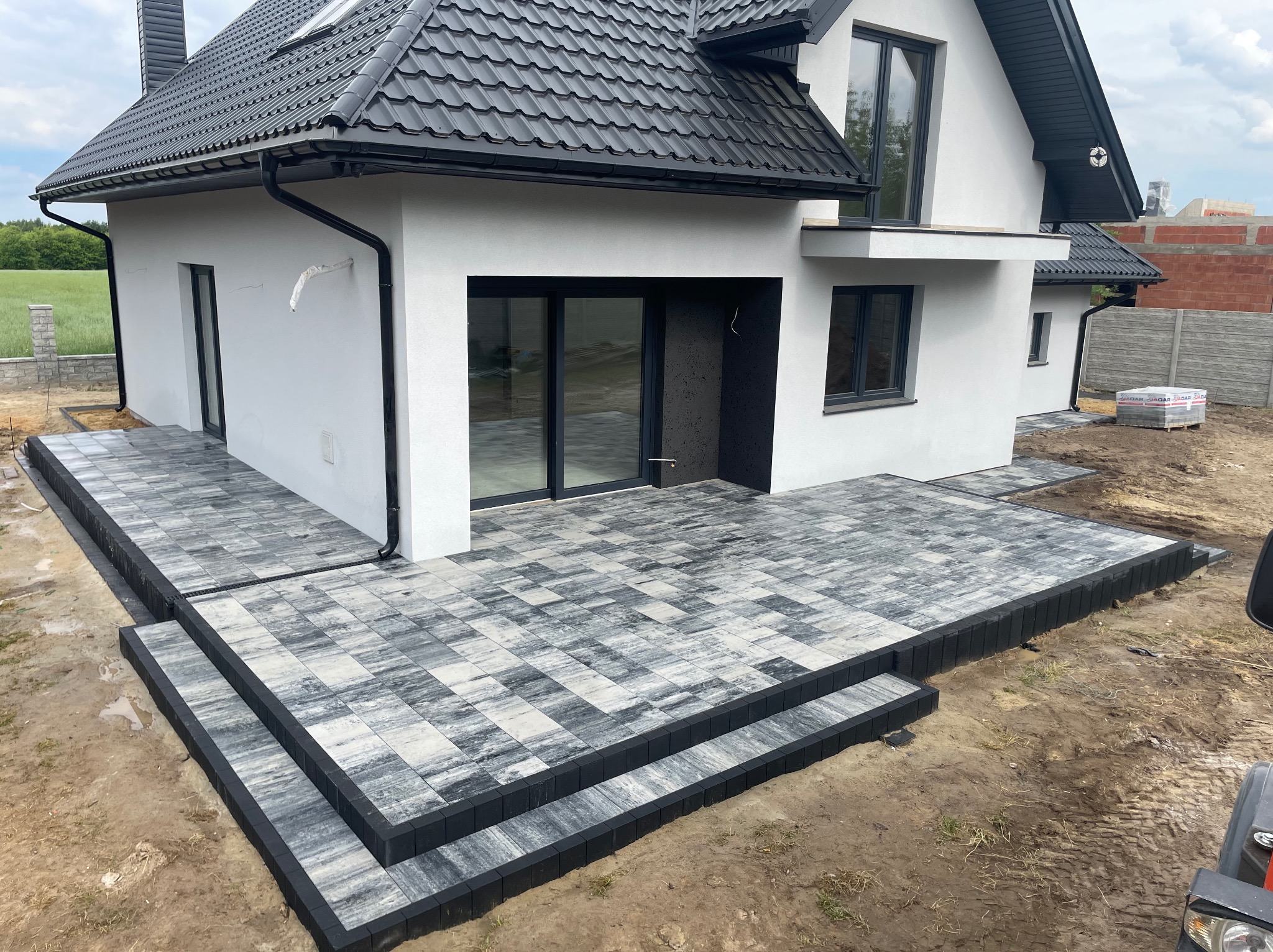 Wykończony taras z kostki brukowej w odcieniach szarości, z czarnym obrzeżem, przylegający do nowoczesnego, białego domu z czarnym dachem i stolarką okienną.