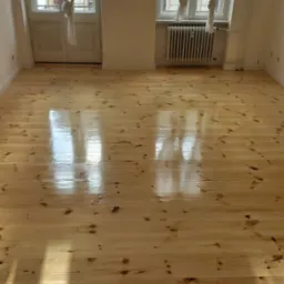 Jasna, odnowiona podłoga z desek w pustym pokoju z dwoma oknami z białymi firankami. Widoczne refleksy światła na lakierowanej powierzchni.