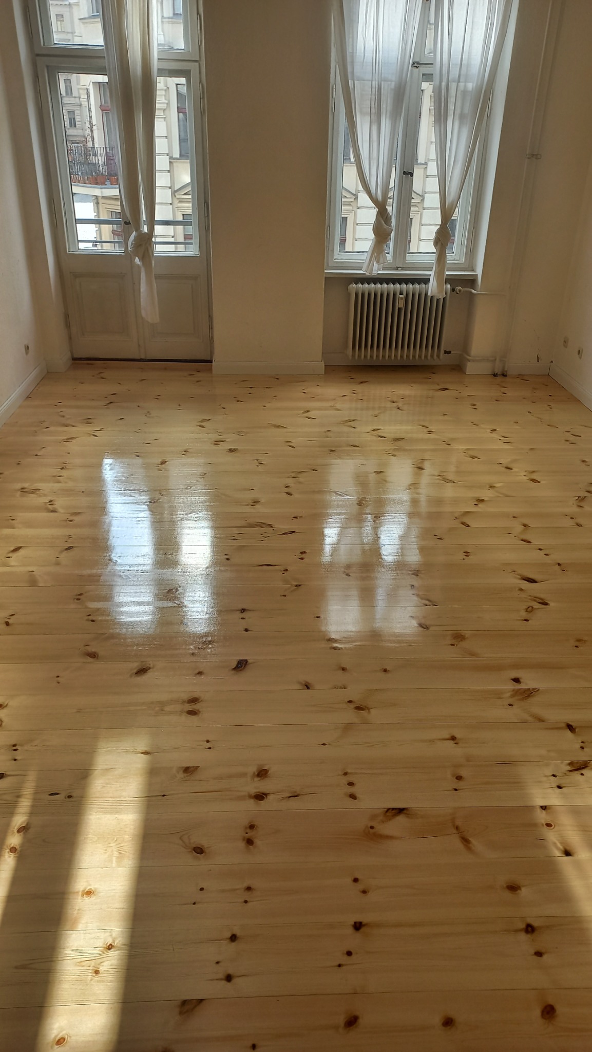 Jasna, odnowiona podłoga z desek w pustym pokoju z dwoma oknami z białymi firankami. Widoczne refleksy światła na lakierowanej powierzchni.