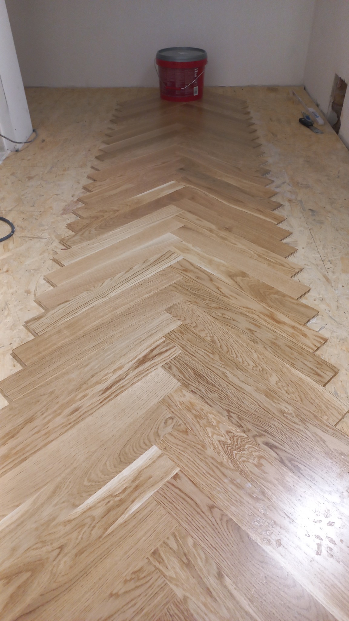 Fragment układania parkietu w jodełkę na podkładzie z płyty OSB, widoczne wiadro z klejem w tle.