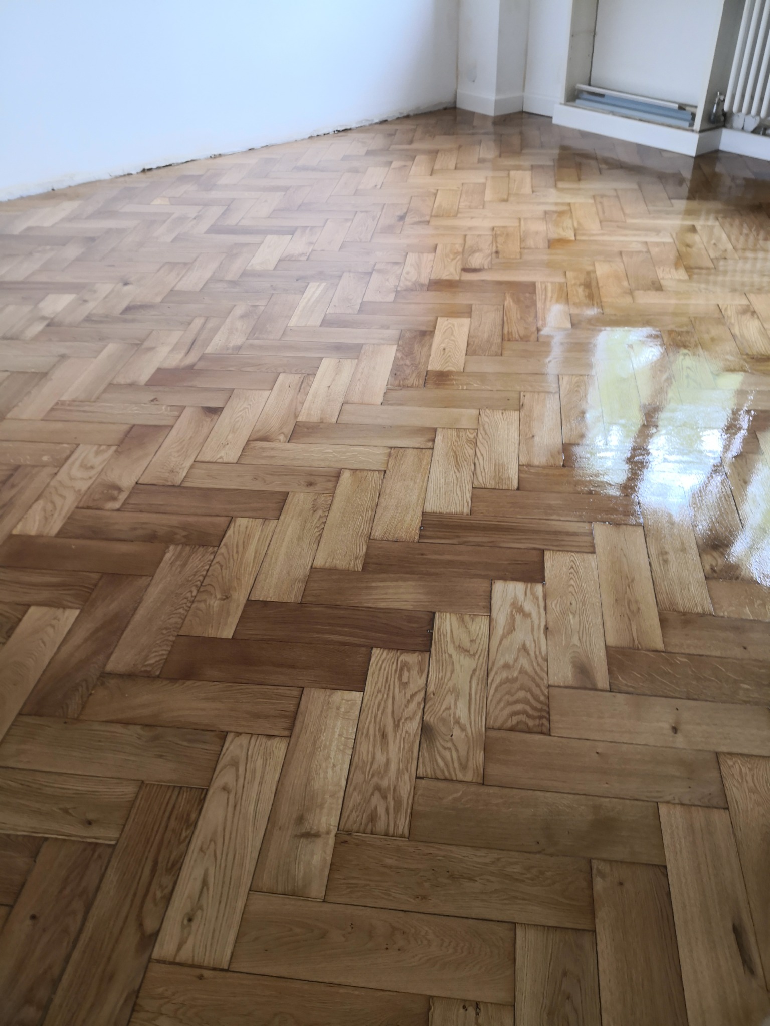 Parkiet ułożony w jodełkę po cyklinowaniu, z widocznym połyskiem świeżo nałożonego lakieru, częściowo jeszcze mokry, odbijający światło okna.