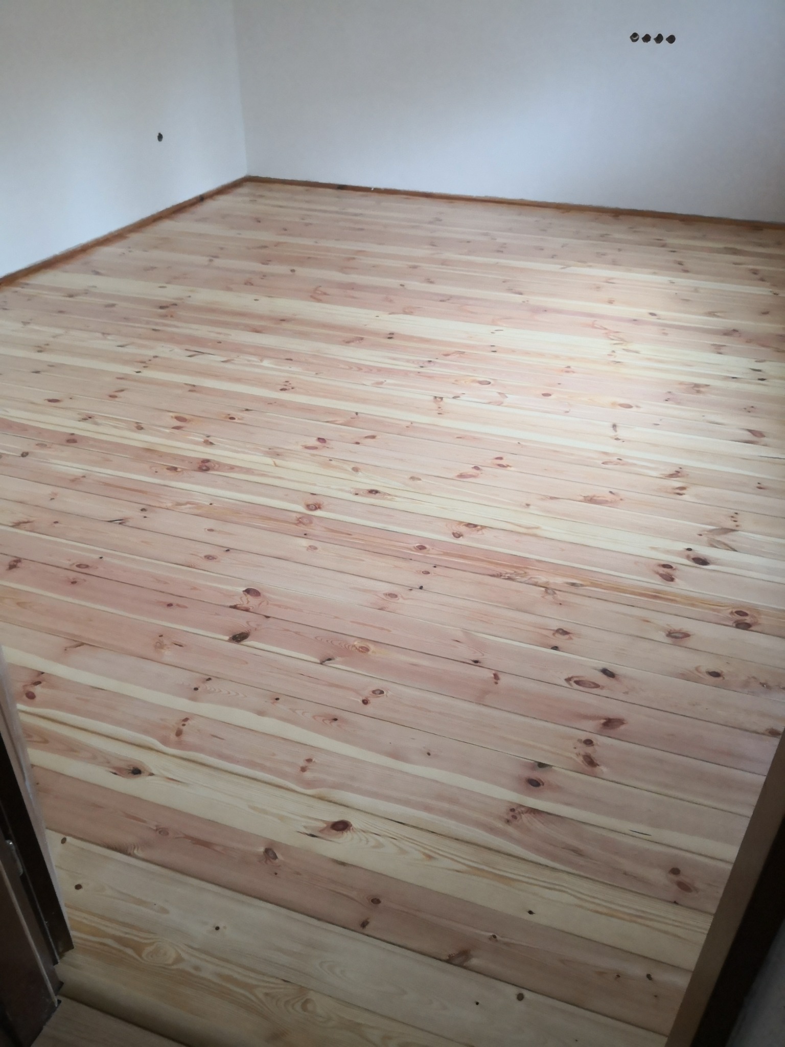 Świeżo położona, jasna podłoga z desek w pustym, jasnym pokoju z widocznymi otworami na kable elektryczne w ścianie.