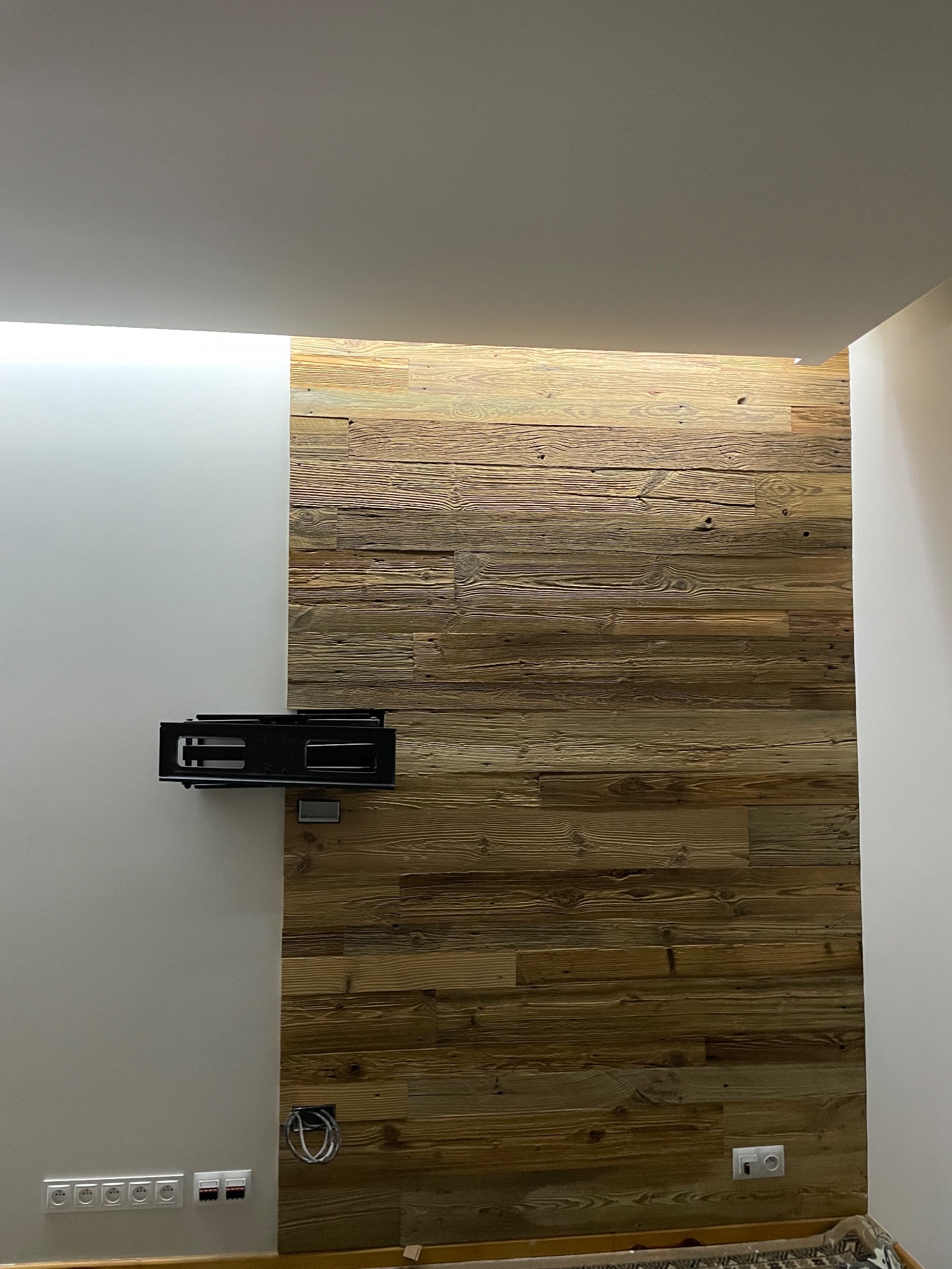 Ściana z paneli drewnianych z zamontowanym uchwytem na telewizor, obok pomalowana ściana z gniazdkami elektrycznymi, oświetlenie sufitowe.