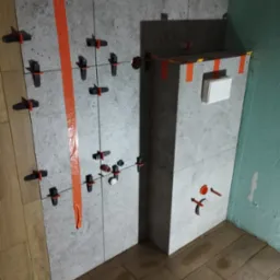 Świeżo ułożone szare płytki z elementami poziomowania, pomarańczową taśmą i widocznymi instalacjami w trakcie remontu łazienki. Na podłodze ułożone płytki drewnopodobne.