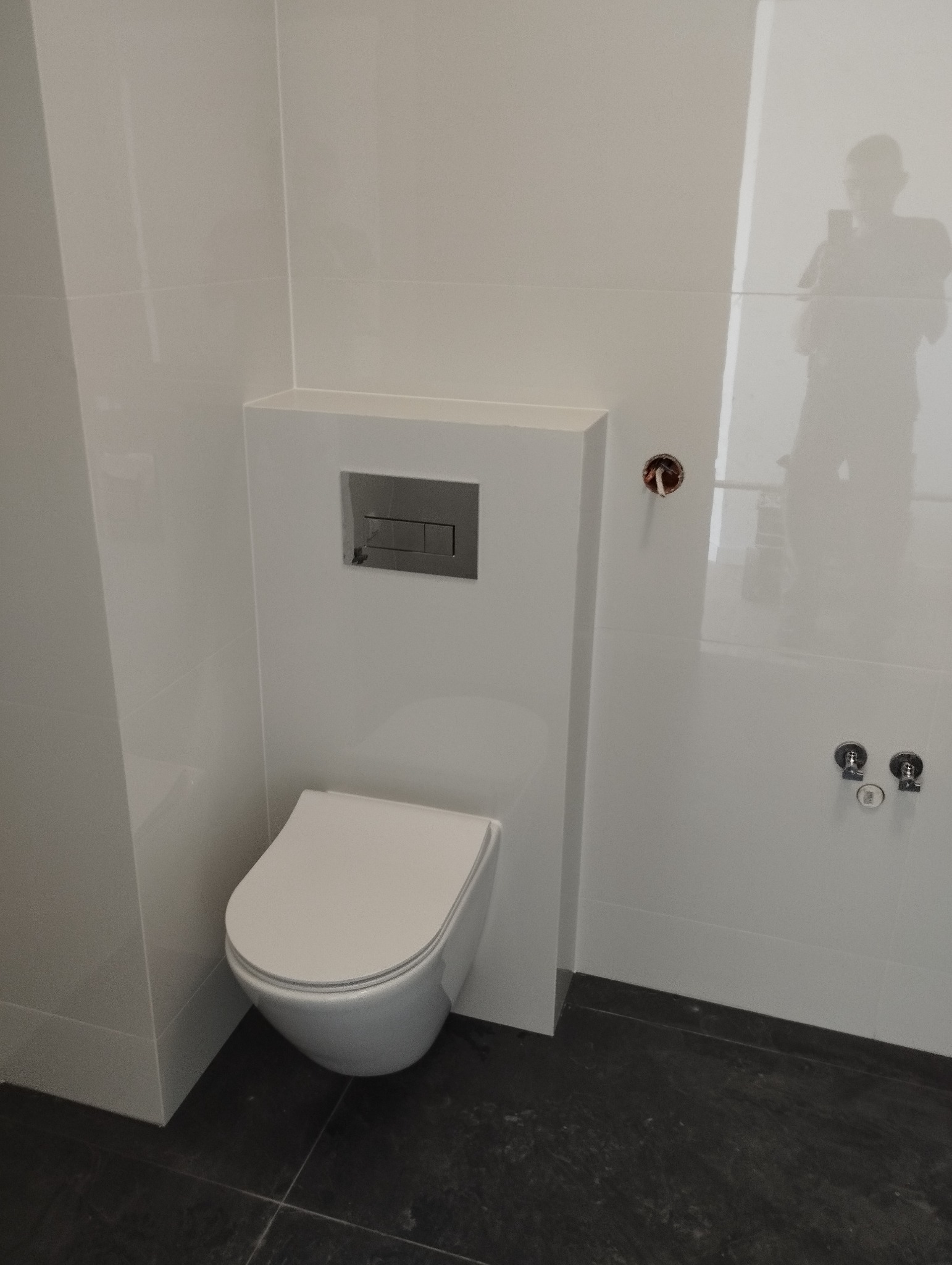Nowoczesna, podwieszana toaleta z chromowanym przyciskiem spłukującym w zabudowie, białe płytki ścienne i ciemna podłoga, widoczne przyłącza wodne i odbicie fotografa w płytkach.