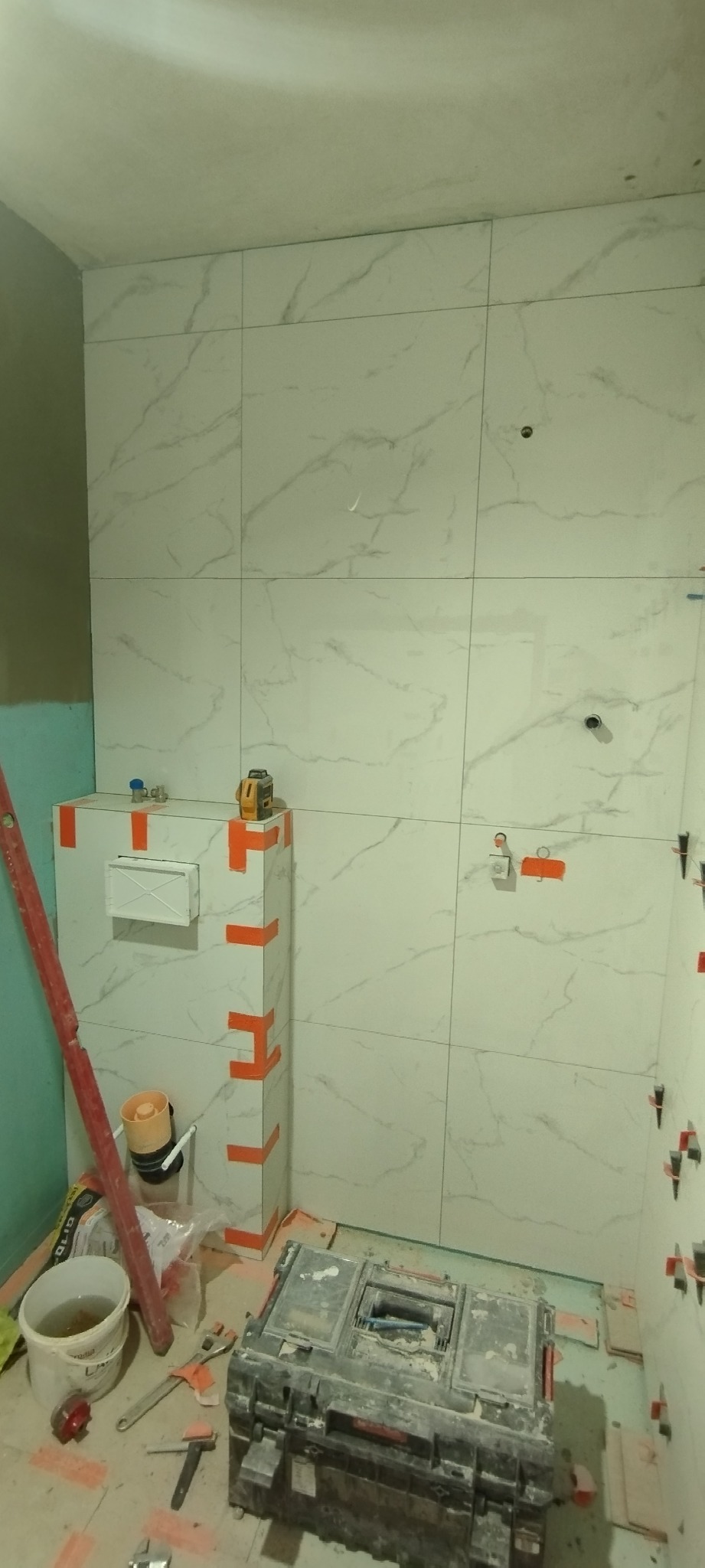 Białe płytki imitujące marmur na ścianie łazienki w trakcie montażu, widoczny poziomica laserowa, skrzynka z narzędziami i akcesoria glazurnicze.