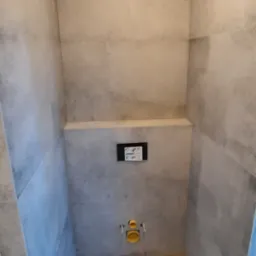 Wnęka w łazience wykończona szarymi płytkami imitującymi beton, z widoczną instalacją podtynkową WC i fragmentem podłogi z paneli imitujących drewno.