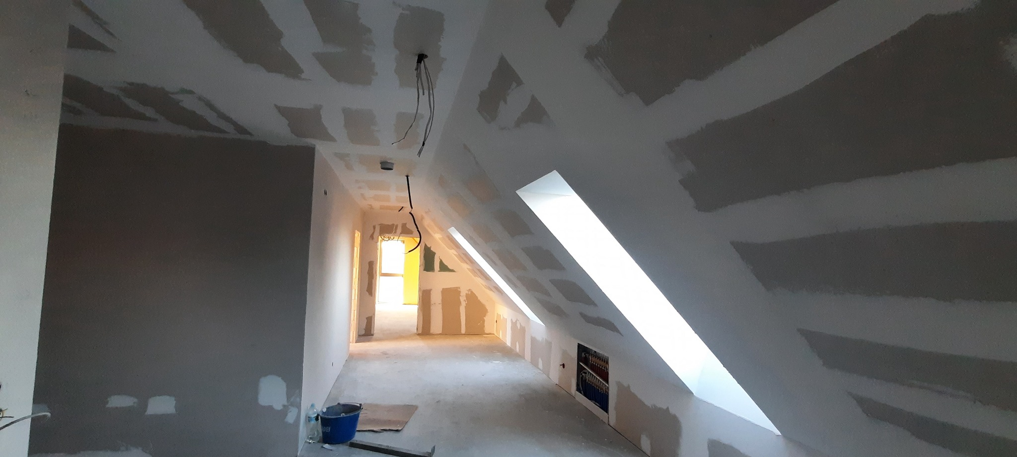 Poddasze w trakcie wykańczania, widoczne płyty kartonowo-gipsowe na ścianach i skosach sufitu, szpachlowane łączenia, wystające przewody elektryczne, okna dachowe wpuszczające jasne światło, wiadro...