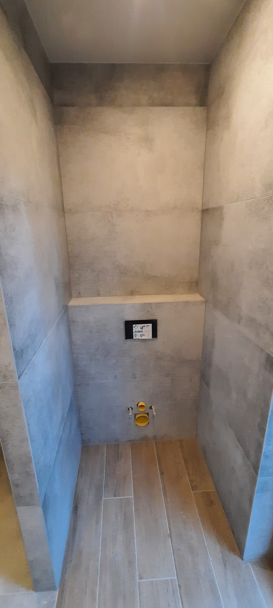 Wnęka w łazience wykończona szarymi płytkami imitującymi beton, z widoczną instalacją podtynkową WC i fragmentem podłogi z paneli imitujących drewno.