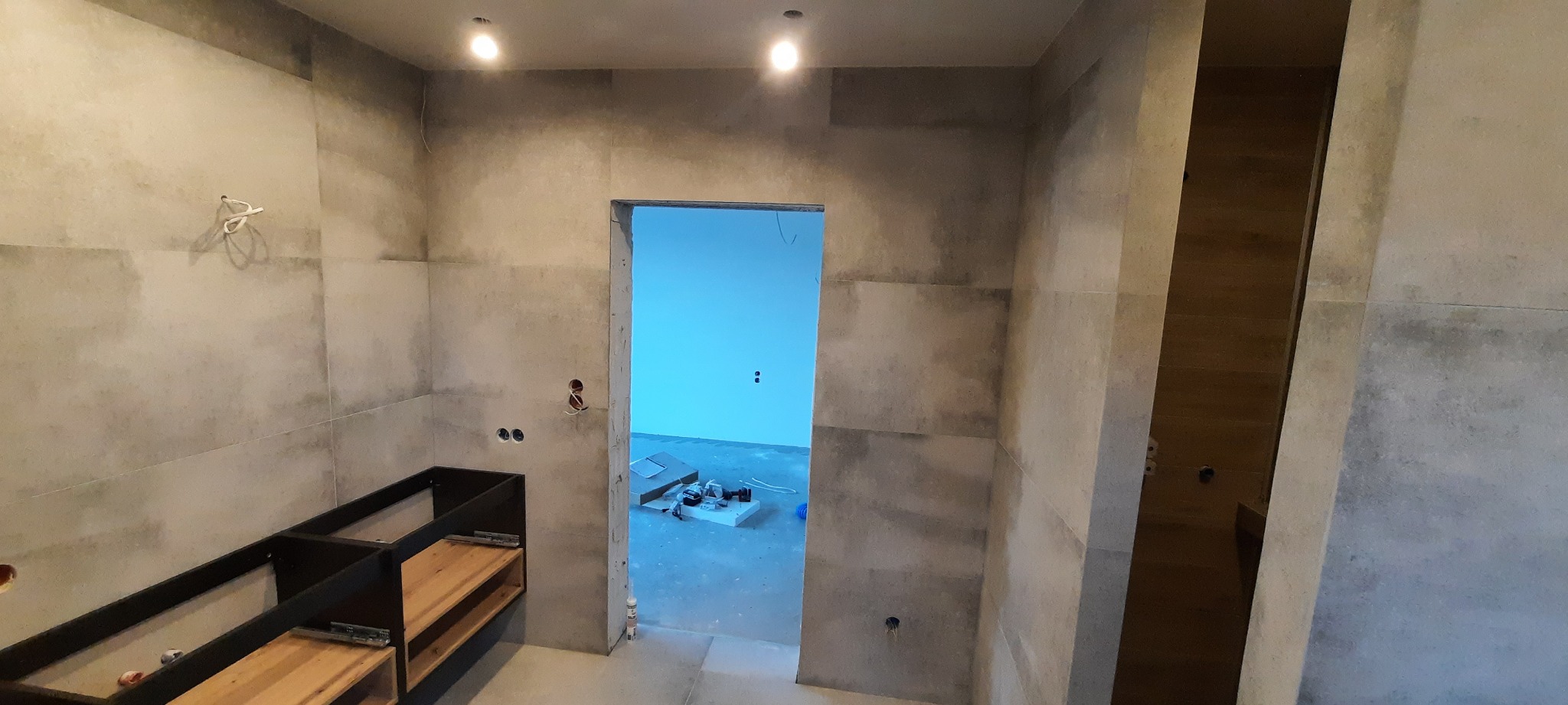 Wnętrze łazienki w trakcie prac wykończeniowych, widoczne szare płytki na ścianach, niedokończona zabudowa szafki pod umywalkę oraz otwór drzwiowy do kolejnego pomieszczenia z niebieskimi ścianami,...