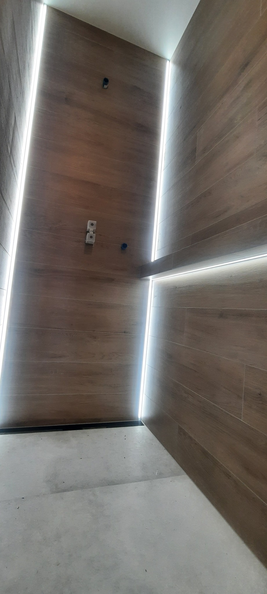 Nowoczesna łazienka z drewnopodobnymi płytkami i oświetleniem LED wzdłuż krawędzi, widoczne gniazdka elektryczne i otwory na armaturę.