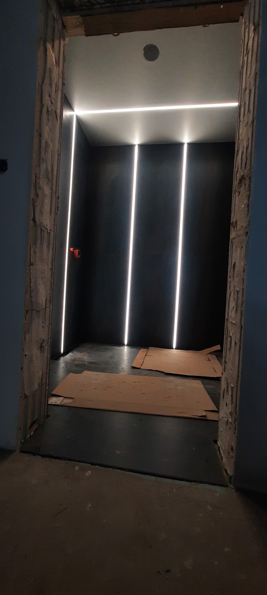 Ciemne pomieszczenie w Górkach Wielkich z zamontowanym oświetleniem LED na ścianach i suficie, zabezpieczone kartonami na podłodze, widok przez surową futrynę drzwiową.